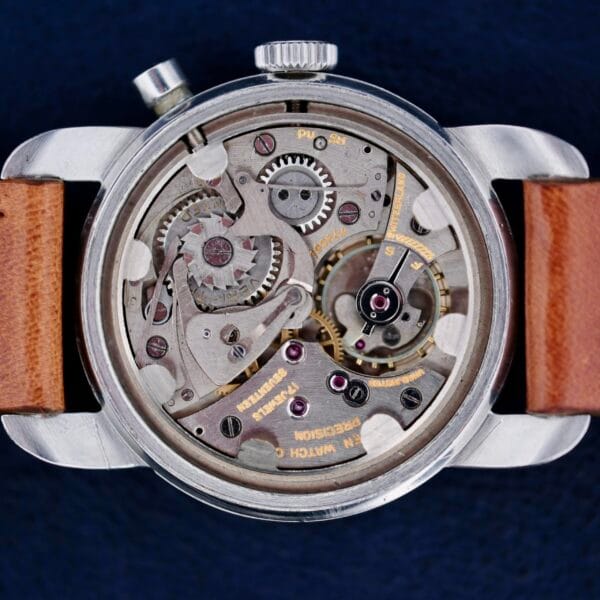 The Gruen Chronotimer Steel with Pulsations Dial showcases a close-up of its open mechanical back, revealing intricate gears and components. It features a silver case and brown leather strap against a dark blue background, accentuating the watchs metallic details.