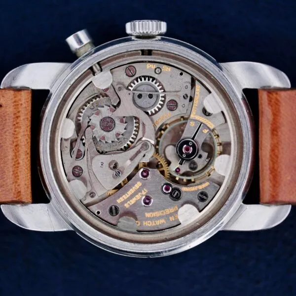 The Gruen Chronotimer Steel with Pulsations Dial showcases a close-up of its open mechanical back, revealing intricate gears and components. It features a silver case and brown leather strap against a dark blue background, accentuating the watchs metallic details.