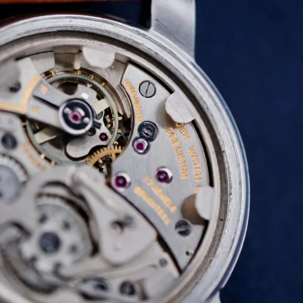 Close-up of a Gruen Chronotimer Steel movement with visible gears, screws, and metal components. WATCH CO and PRECISION are engraved. The design showcases intricate mechanical parts interlocking against a dark blue background.