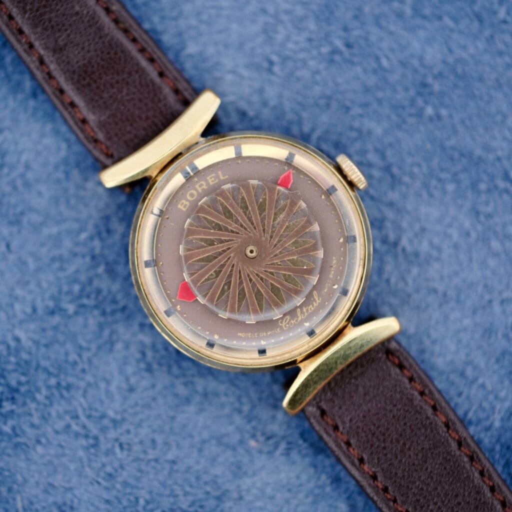 An Ernest Borel Kaleidoscope Cocktail Brown Dial watch with a gold case sits on a blue textured surface.
