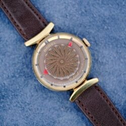 An Ernest Borel Kaleidoscope Cocktail Brown Dial watch with a gold case sits on a blue textured surface.