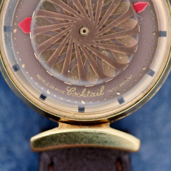 Close-up of an Ernest Borel Kaleidoscope Cocktail wristwatch with a gold-toned case, brown patterned dial, red hour markers, and Modèle Déposé Swiss Made inscription. It features a leather strap.