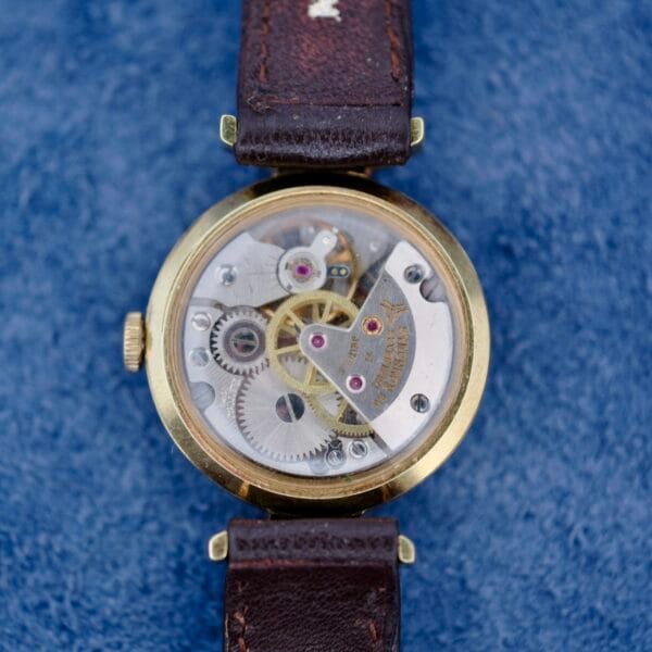 A close-up of the Ernest Borel Kaleidoscope Cocktail Brown Dial watch reveals intricate gears through its transparent case. Featuring a brown leather strap, it rests on a blue textured surface.