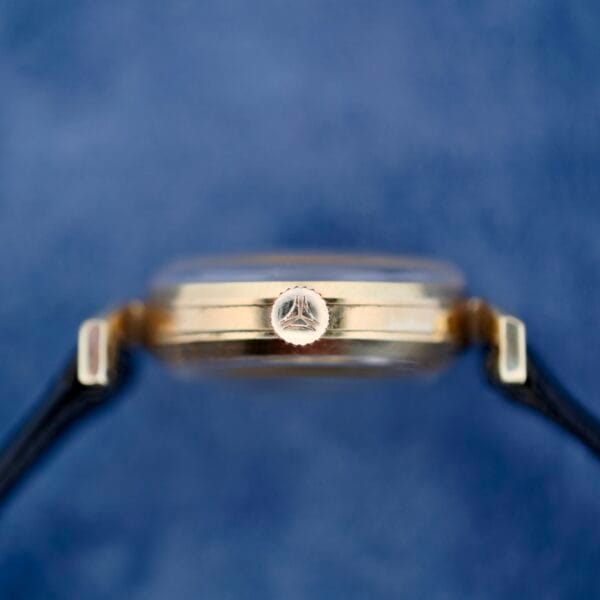 Side view of the Ernest Borel Kaleidoscope Cocktail with a focus on its triangular logo-adorned crown, set against a blurred blue background. The watch features a gold-toned case and a partially visible black leather strap.