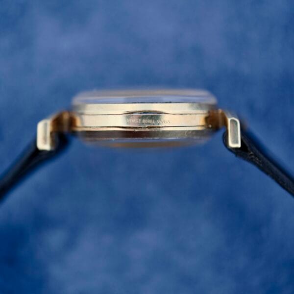 Side view of an Ernest Borel Kaleidoscope Cocktail wristwatch with a thin metallic case engraved “Lynvet Forsey Swiss,” set against a blue background. The watch features black leather straps and a clear glass face.