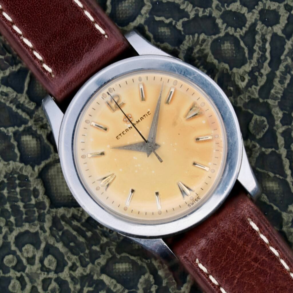 Eterna-Matic Tropic Dial Hidden Crown watch with a brown leather strap, showcased on a textured dark background.