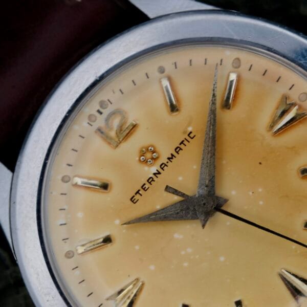Close-up of a vintage Eterna-Matic Tropic Dial Hidden Crown wristwatch featuring a beige face with metallic details, hour, minute, and second hands. The center displays Eterna-Matic, and it is paired with a brown leather band.