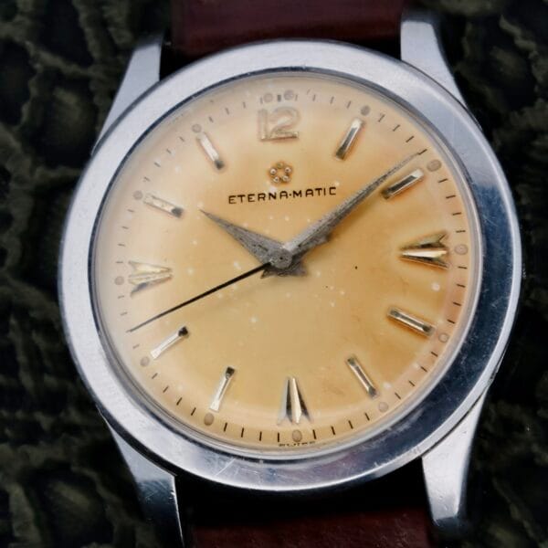 A vintage Eterna-Matic Tropic Dial Hidden Crown wristwatch with a yellowed dial, silver hands and hour markers, Roman numerals at 12, 3, 6, and 9 positions, a silver case, and a brown leather strap.