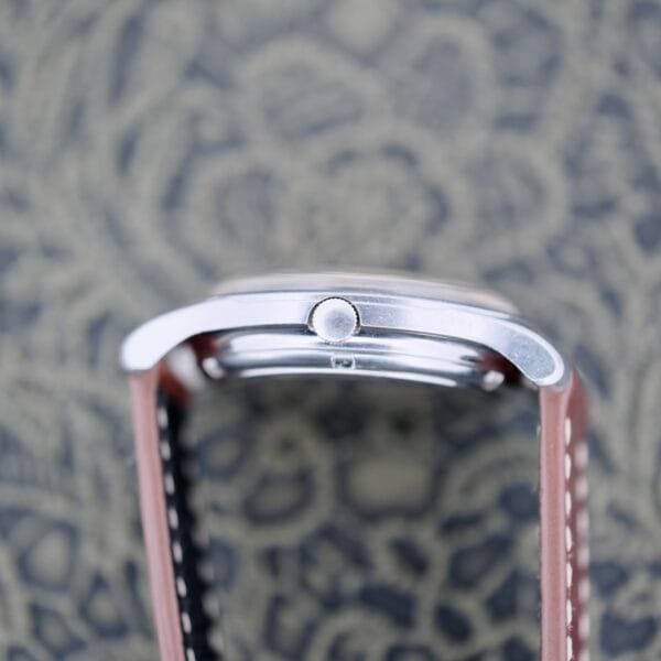 The Eterna-Matic Tropic Dial Hidden Crown wristwatch, with its sleek silver case and round crown, rests on a blurred patterned surface. It features a brown leather strap that complements the intricate backdrop.