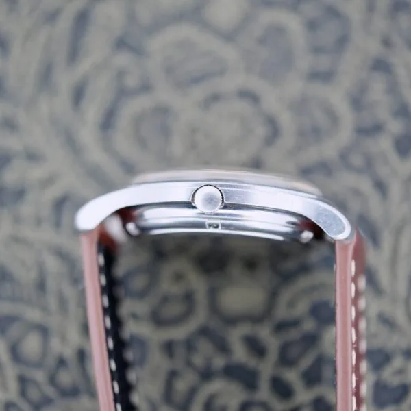 The Eterna-Matic Tropic Dial Hidden Crown wristwatch, with its sleek silver case and round crown, rests on a blurred patterned surface. It features a brown leather strap that complements the intricate backdrop.
