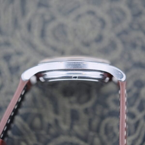 Side view of the Eterna-Matic Tropic Dial Hidden Crown watch with a silver metal frame and a brown leather strap, set against a backdrop of blurred black and gray patterned fabric.