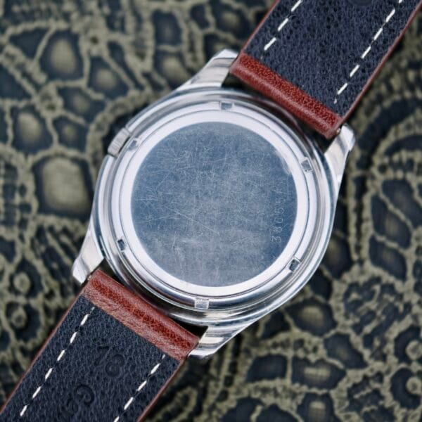 Close-up of the back of an Eterna-Matic Tropic Dial Hidden Crown wristwatch with a scratched stainless steel case. The brown and black leather strap, stitched with white thread, is visible as it rests on a patterned fabric surface.