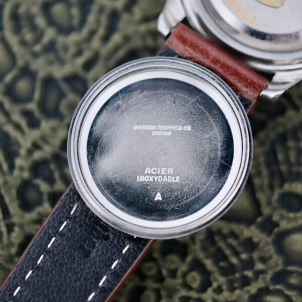 Close-up of an Eterna-Matic Tropic Dial Hidden Crown watch with the back cover removed, revealing text such as SWISS MADE, ACIER INOXYDABLE, and numbers. It features a brown leather strap with white stitching against a textured background.