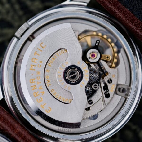 Close-up of a mechanical watch movement with visible metal components, gears, and inscriptions. The text reads Eterna-Matic and highlights features like a rotor and jeweled bearings. A leather strap partially frames the Eterna-Matic Tropic Dial Hidden Crown watch image.