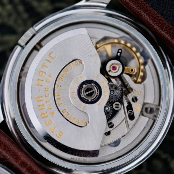 Close-up of a mechanical watch movement with visible metal components, gears, and inscriptions. The text reads Eterna-Matic and highlights features like a rotor and jeweled bearings. A leather strap partially frames the Eterna-Matic Tropic Dial Hidden Crown watch image.