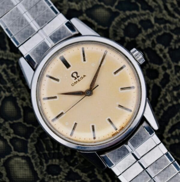 The Omega Lemon Tropic Dial 14390 Stainless Steel wristwatch features a classic round cream face with silver hour markers and the iconic Omega logo under 12 oclock. Its sleek silver metal bracelet stands out against a textured dark green fabric backdrop, capturing timeless elegance.