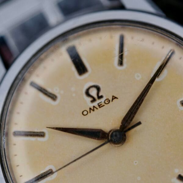 Close-up of a vintage Omega Lemon Tropic Dial 14390 wristwatch face, showcasing the iconic logo. The cream-colored dial features black hour markers and silver hands, displaying its history with scratches and patina.