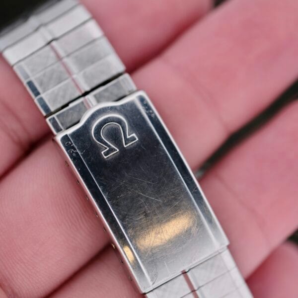 Close-up of a hand holding a clasp showcasing the Omega Lemon Tropic Dial 14390. The stainless steel wristband has light scratches, a brushed finish, and a sophisticated linked design that embodies Omegas timeless elegance, complete with the iconic logo.