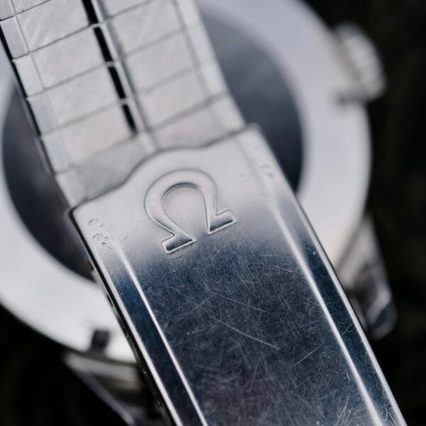 Close-up of the Omega Lemon Tropic Dial 14390 Stainless Steel strap with a metal clasp featuring the iconic logo, showing character-revealing scratches. The watch case and crown subtly peek through in the background, highlighting its timeless elegance.