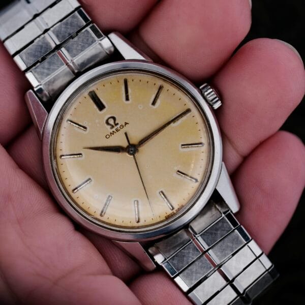 A hand holds the Omega Lemon Tropic Dial 14390, showcasing its beige face with simple hour markers and two hands. The iconic Omega logo adorns the dial, while the gleaming stainless steel casing enhances its vintage allure.