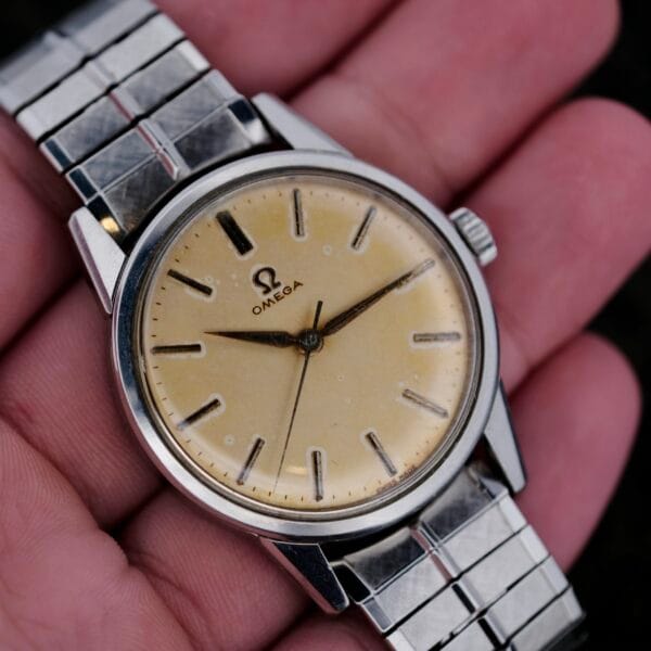 A hand showcases the Omega Lemon Tropic Dial 14390, featuring a round beige dial, slightly worn. Its silver stainless steel band complements minimalist hour markers and two hands, with the Omega logo elegantly beneath the 12 oclock mark.