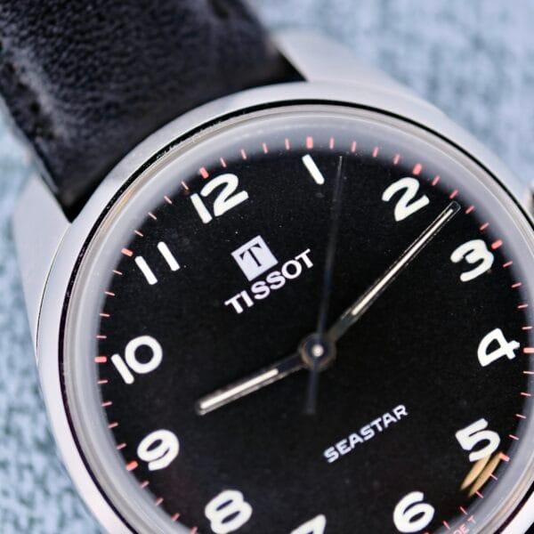 Close-up of a Tissot Seastar Manual Wind Arabic Numerals wristwatch with a black leather strap, featuring a black face, silver hour and minute hands, and white numerals. The TISSOT brand name is visible near the 12 oclock position.