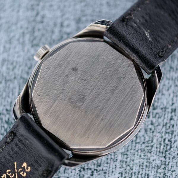 The image displays the back of a Tissot Seastar Manual Wind Arabic Numerals wristwatch with a brushed metal case. Its black leather strap, partially visible, shows wear and white markings against a textured gray background.
