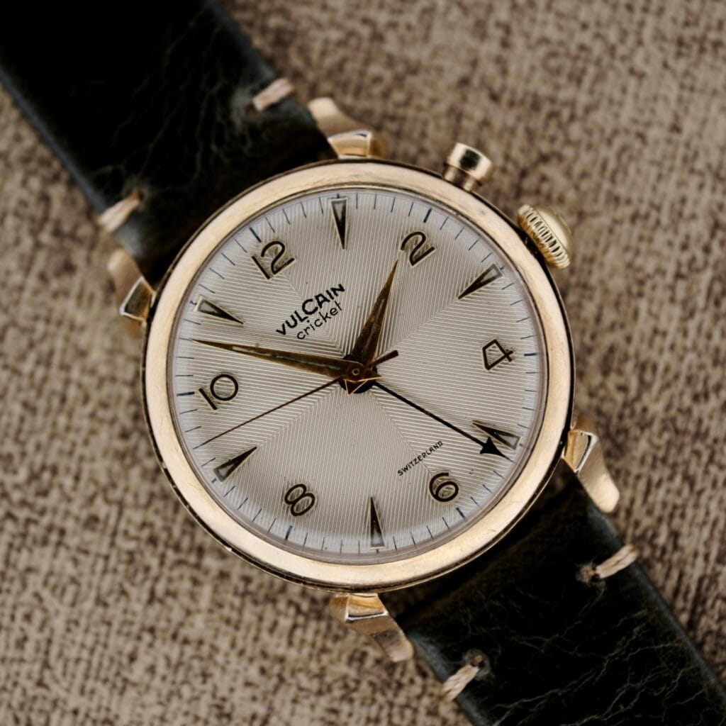 A Vulcain Cricket 120 Radial Dial Crab Lugs wristwatch with distinctive crab lugs and a black leather strap rests elegantly on a textured surface.