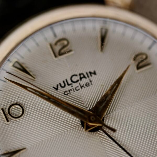 Close-up of a Vulcain Cricket 120 Radial Dial Crab Lugs watch face, featuring a textured white dial with gold, pointed hour markers and hands. The brand name VULCAIN and cricket are printed near the center. The time reads approximately 10:10.