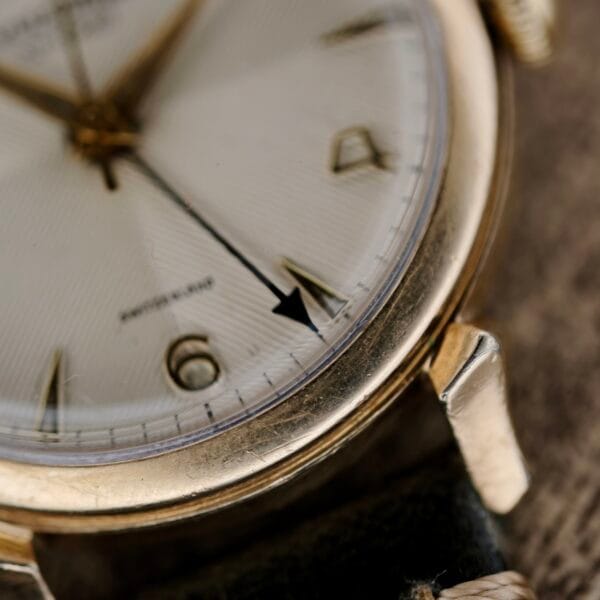 Close-up of a Vulcain Cricket 120 Radial Dial Crab Lugs, a vintage gold wristwatch with a white dial and black leather strap. The watchs hands point near 4:20, showing numerals at 4 and 6 with simple line markers, and displays some signs of wear.