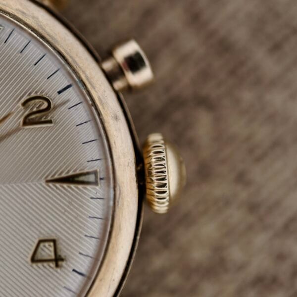 Close-up of the Vulcain Cricket 120 Radial Dial Crab Lugs wristwatch with metallic casing, showcasing a textured silver dial displaying numbers 2 and 4. The crown stands out against a blurred fabric backdrop.
