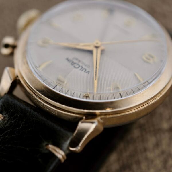 Close-up of a Vulcain Cricket 120 Radial Dial wristwatch with a gold case, textured white dial, gold indices, and black leather strap. The crown and crab lugs are visible, with the brand name partially seen on the dial.