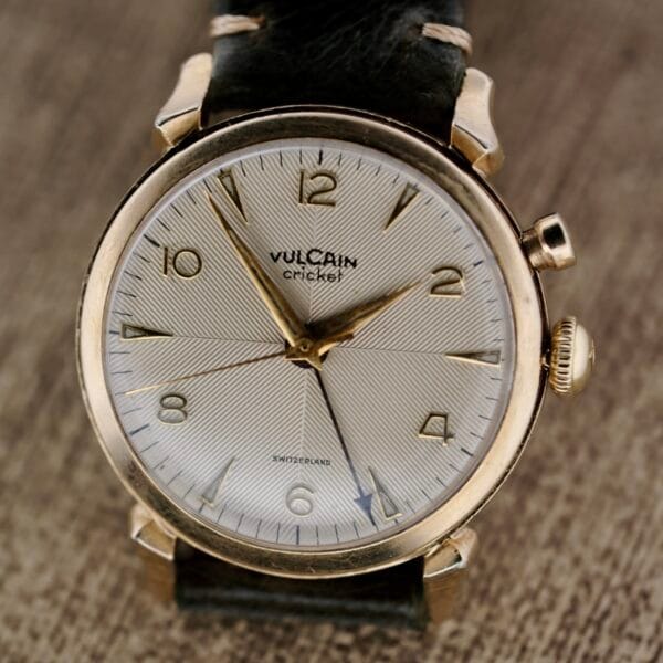 The Vulcain Cricket 120 Radial Dial Crab Lugs is a vintage wristwatch with a textured silver dial, gold hour markers, and hands. It features a crown and pusher on the right side and comes with a black leather strap, displayed on a wooden surface.