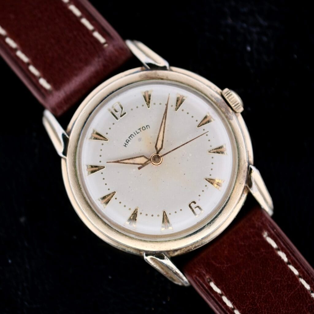 Hamilton Manual Wind Gold Tone wristwatch featuring a gold tone case and brown leather strap, manual wind movement, showcased on a black background.