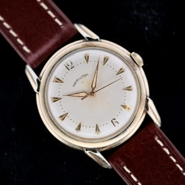 The Hamilton Manual Wind Gold Tone wristwatch features a round white dial with silver triangular hour markers and two hands, encased in metallic gold tone and paired with a brown leather strap. It is displayed on a black background.