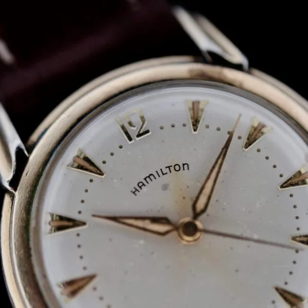 Close-up of a vintage Hamilton Manual Wind Gold Tone wristwatch with silver and gold tones. The dial features pointed gold hour markers, two hands, and the brand name Hamilton at the center. The watch face shows signs of wear against a dark background.