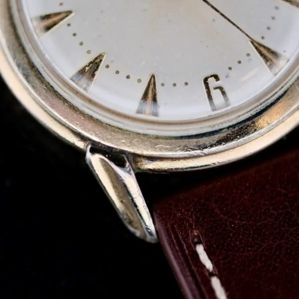 Close-up of the Hamilton Manual Wind Gold Tone watch face displaying hour markers and the number six. The silver case pairs with a brown leather strap featuring visible stitching, set against a dark background to highlight the watchs details.
