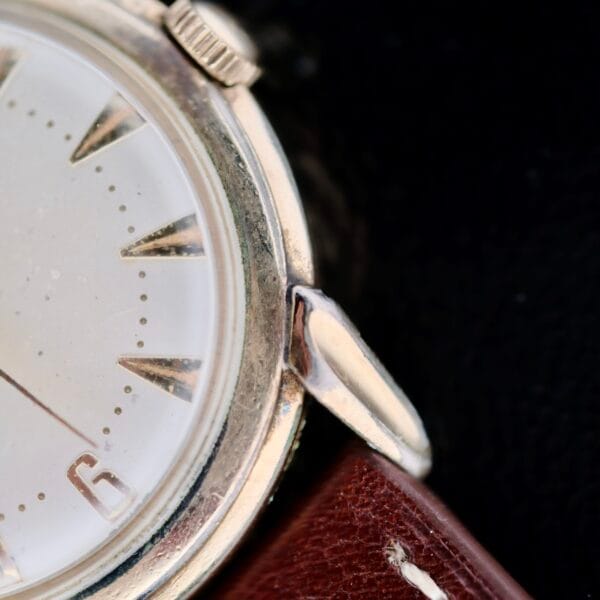 A close-up of the Hamilton Manual Wind Gold Tone wristwatch reveals a vintage round silver bezel, a minimalist white dial with triangular hour markers, and part of the number six. The watch is paired with a brown leather strap.