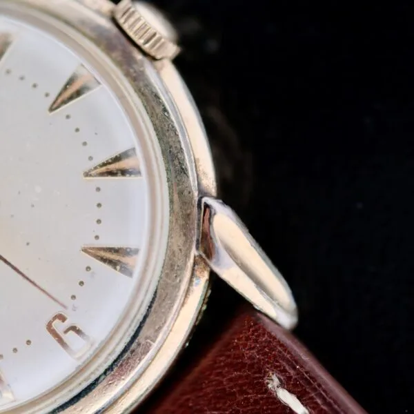 A close-up of the Hamilton Manual Wind Gold Tone wristwatch reveals a vintage round silver bezel, a minimalist white dial with triangular hour markers, and part of the number six. The watch is paired with a brown leather strap.