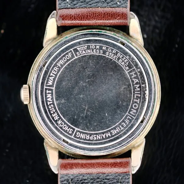 The image reveals the back of a Hamilton Manual Wind Gold Tone wristwatch with a brown and black leather strap. Inscribed on the case are: Hamilton, Stainless Steel Back, 10K R.G. Bezel, WaterProof, Shock-Resistant, Anti-Magnetic, Lifetime Mainspring. The background is black.