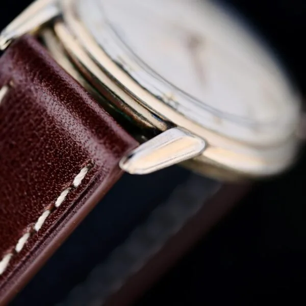 The Hamilton Manual Wind Gold Tone watch features a brown leather strap with white stitching, a metallic case with visible lugs, and an out-of-focus dial set against a dark background highlighting its details.