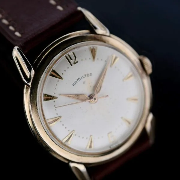 Close-up of a Hamilton Manual Wind Gold Tone watch with a round, gold-toned case and a white dial featuring gold hour markers and hands, set against a dark brown leather strap, photographed on a black background.