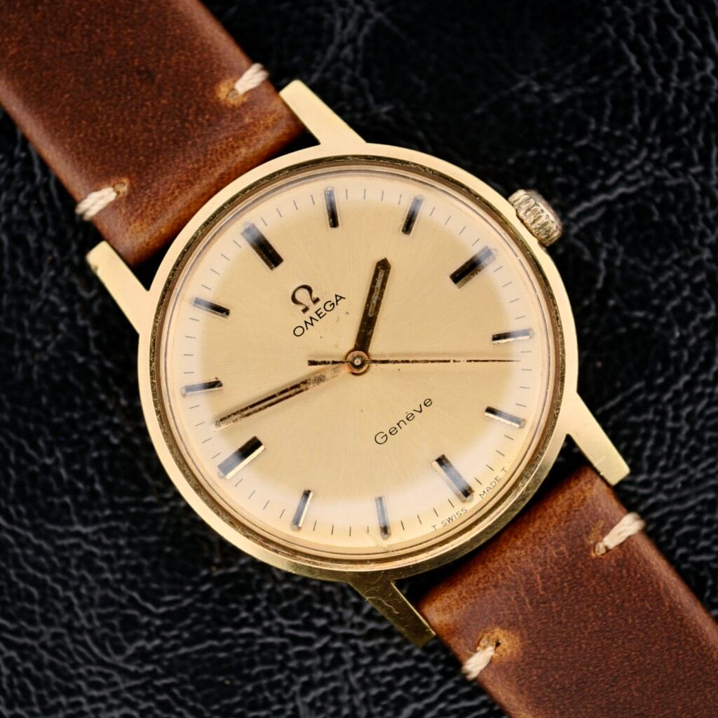 Omega Geneve Gold Champagne Dial 135.070 watch with a brown leather strap, displayed on a black textured surface.