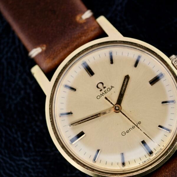 An Omega Geneve Gold Champagne Dial 135.070 wristwatch with a gold-toned case features a beige dial, black hour markers, and hands. A brown leather strap complements its design, with the Omega logo and Genève clearly visible on the dial.