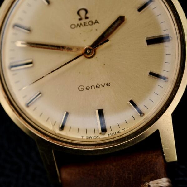 Close-up of an Omega Geneve Gold Champagne Dial 135.070 watch with a beige dial and black hour markers. It features a brown leather strap, Swiss Made at the bottom, and slight wear on the face.