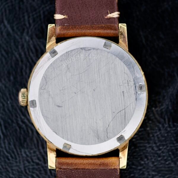 The image depicts the back of an Omega Geneve Gold Champagne Dial 135.070 watch, featuring a worn silver-toned case and a brown leather strap with minor scratches on the metal. It rests on a textured black surface.