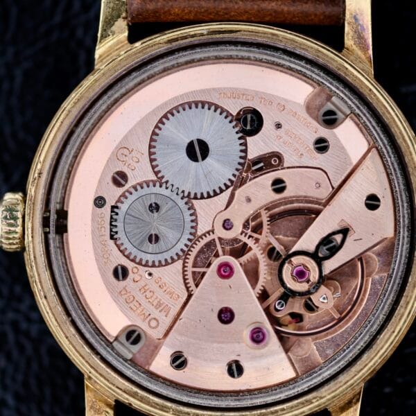 A close-up view of the Omega Geneve Gold Champagne Dial 135.070 reveals its intricate movement with gears, cogwheels, and screws elegantly arranged within a gold-colored casing featuring visible engravings.