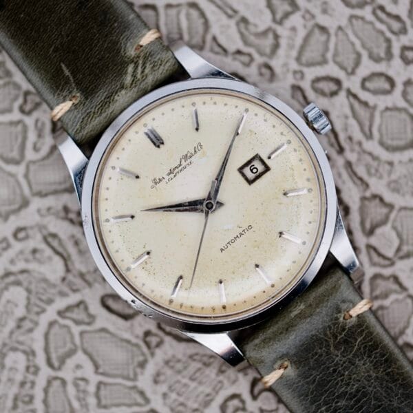 The IWC Stainless Steel Automatic C.853 watch is showcased, featuring a cream dial with minimal hour markers, a date window at three oclock, and a silver casing. It is paired with a worn dark green leather strap against a textured patterned backdrop.