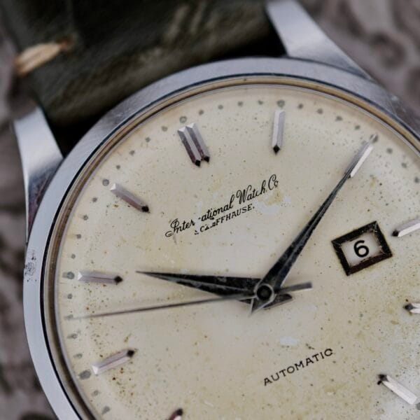 Close-up of the IWC Stainless Steel Automatic C.853 wristwatch featuring a cream-colored face, silver hour markers, and hands. The date shows 6, and there are signs of aging like speckles and wear. It has the inscription International Watch Co. Schaffhausen.