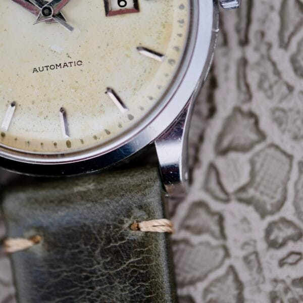 Close-up of an IWC Stainless Steel Automatic C.853 wristwatch featuring a cream face, round silver bezel, and black markers. The watch displays AUTOMATIC and a partial date function. It comes with a distressed green leather strap against a textured backdrop.